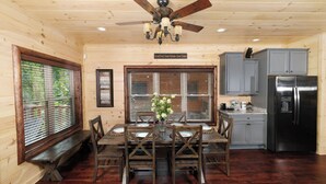 A wooden cabin interior with a dining table, chairs, a ceiling fan, a refrigerator, and a window with blinds. - Quittin Time - Hot Tub, Game Room & Home Theater (Gatlinburg)