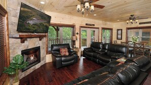 A spacious living room with leather sofas, a fireplace, and a large TV mounted on a stone wall. - Quittin Time - Hot Tub, Game Room & Home Theater (Gatlinburg)