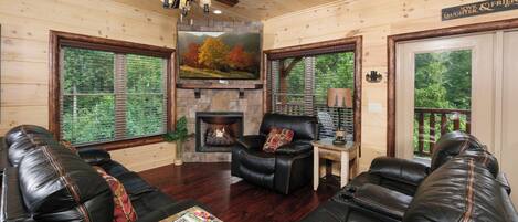A cozy cabin interior with a fireplace, leather sofas, a coffee table with magazines, and large windows offering a view of the forest.