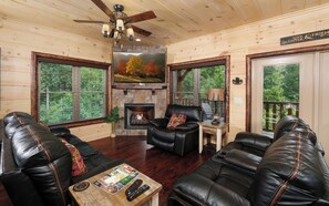 A cozy cabin interior with a fireplace, leather sofas, a coffee table with magazines, and large windows offering a view of the forest.
