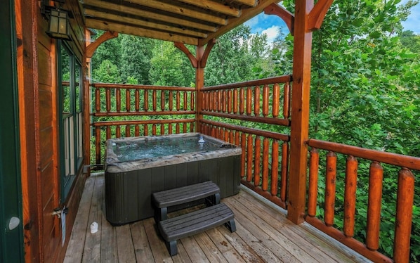 A wooden deck with a hot tub and a bench, surrounded by a wooden railing and lush greenery.