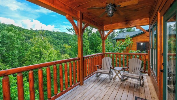 A wooden deck with two Adirondack chairs and a table, overlooking a forested area. - Quittin Time - Hot Tub, Game Room & Home Theater (Gatlinburg)