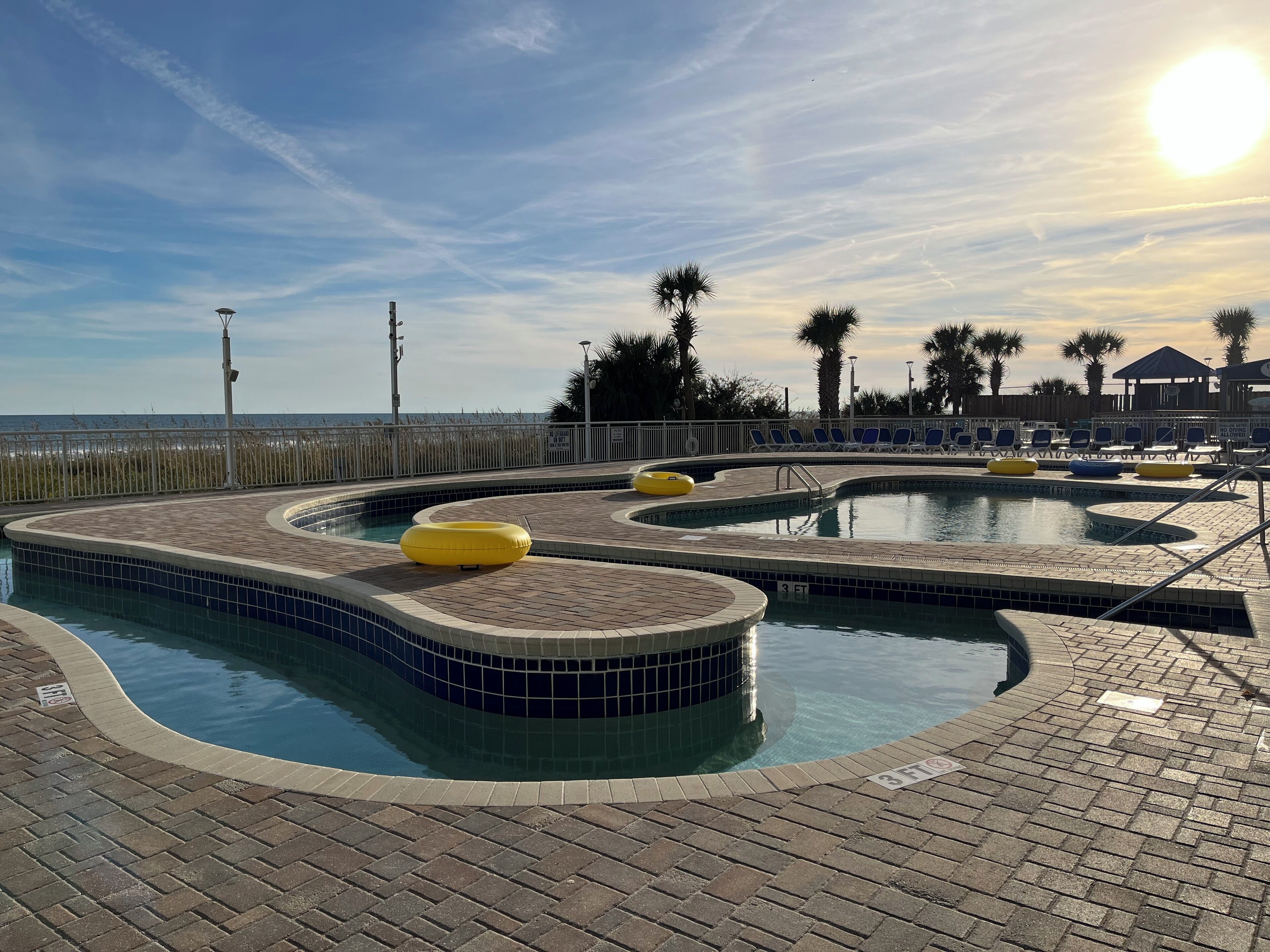 A pool area with a tiled deck, lounge chairs, and a view of the beach.