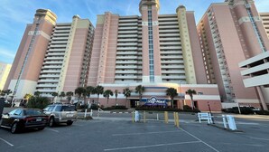 Un hotel de varios pisos con un llamativo letrero de 'Bay Watch', un estacionamiento con varios coches y palmeras en primer plano.