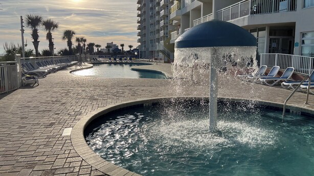 Zona de piscina con fuente, cielo iluminado por el sol y sillas de descanso.