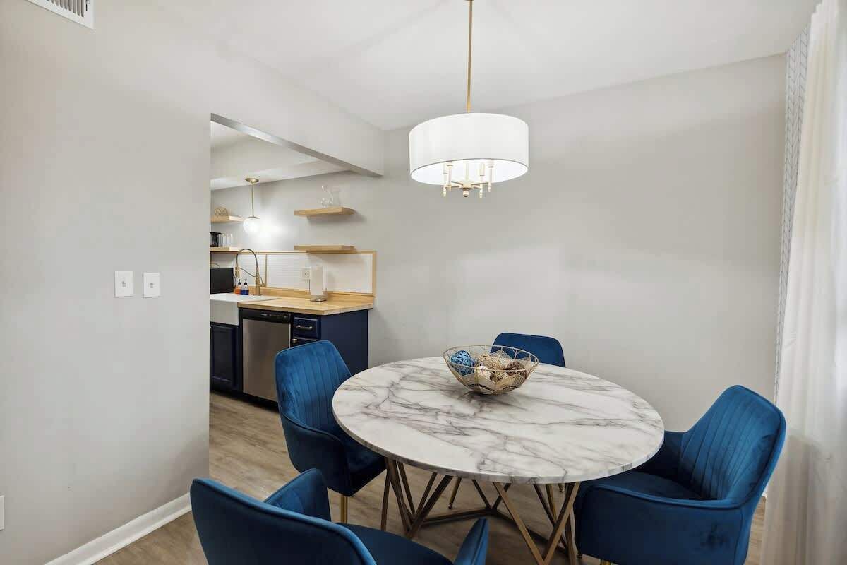 A dining area with a round marble table, blue chairs, a chandelier, and a kitchen in the background.