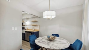 Een eetgedeelte met een ronde marmeren tafel, blauwe stoelen, een kroonluchter en een keuken op de achtergrond.