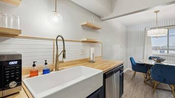 A modern kitchen with a white sink, wooden countertop, and a built-in dishwasher. There is a dining area with a round table and blue chairs.