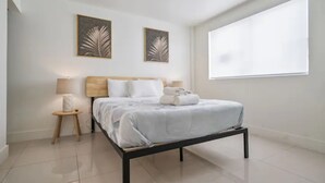 A modern bedroom with a bed, two wall-mounted lamps, a small side table, and a large window. - Miami Suite 203 (Miami)