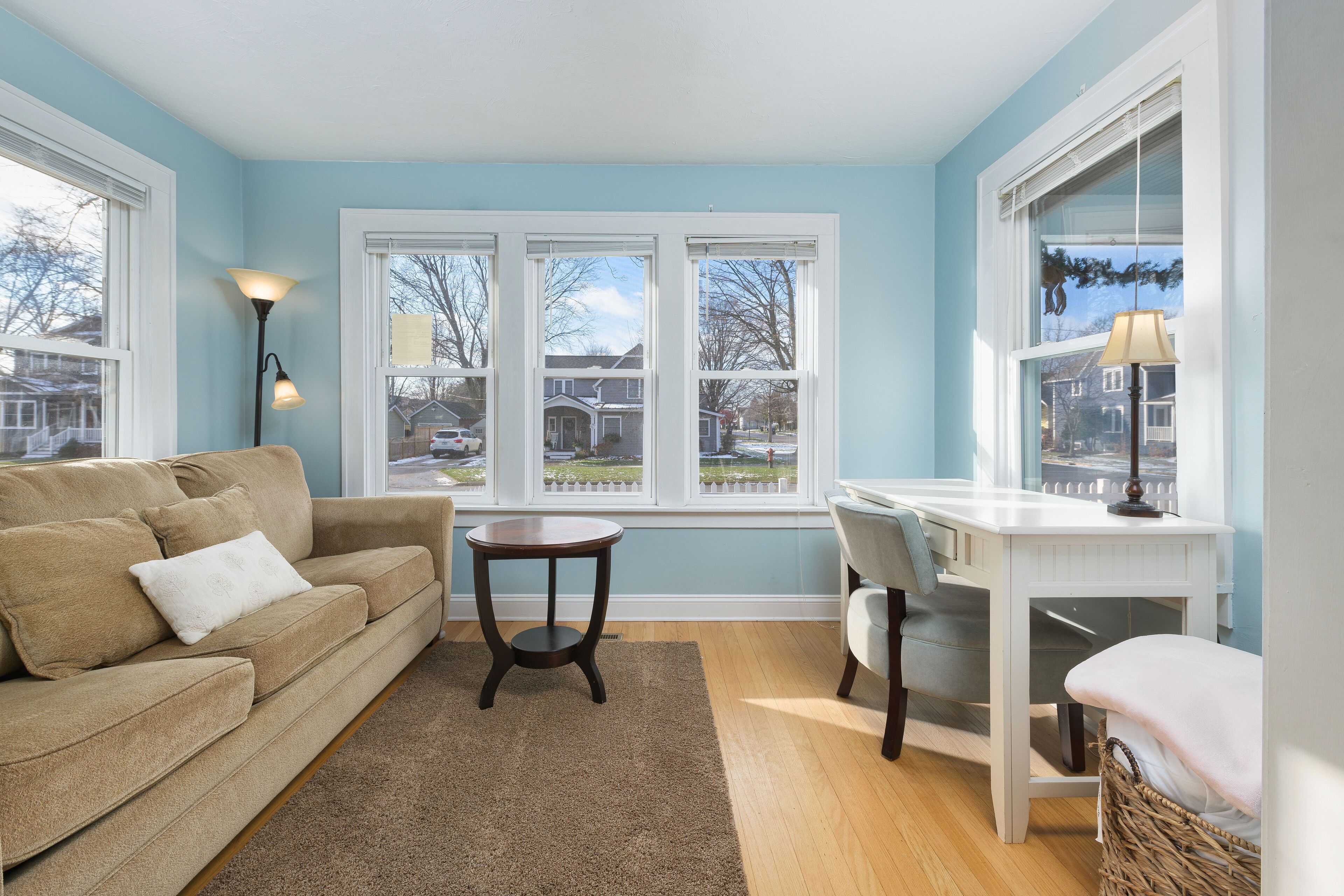 A living room with a beige sofa, a round coffee table, and a white desk with a chair. There are large windows with a view of houses and trees.