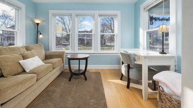 A living room with a beige sofa, a round coffee table, and a white desk with a chair. There are large windows with a view of houses and trees.