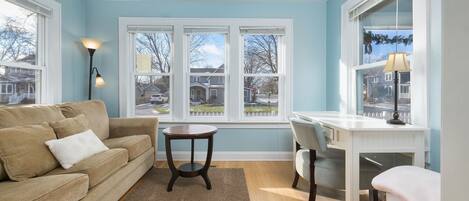 Un salon avec un canapé beige, une table basse ronde et un bureau blanc avec une chaise. De grandes fenêtres offrent une vue sur les maisons et les arbres.