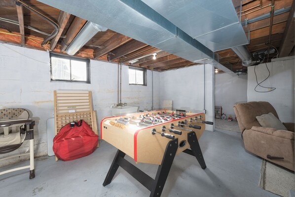 A room with a foosball table, a red bag, a wooden chair, and a sofa.