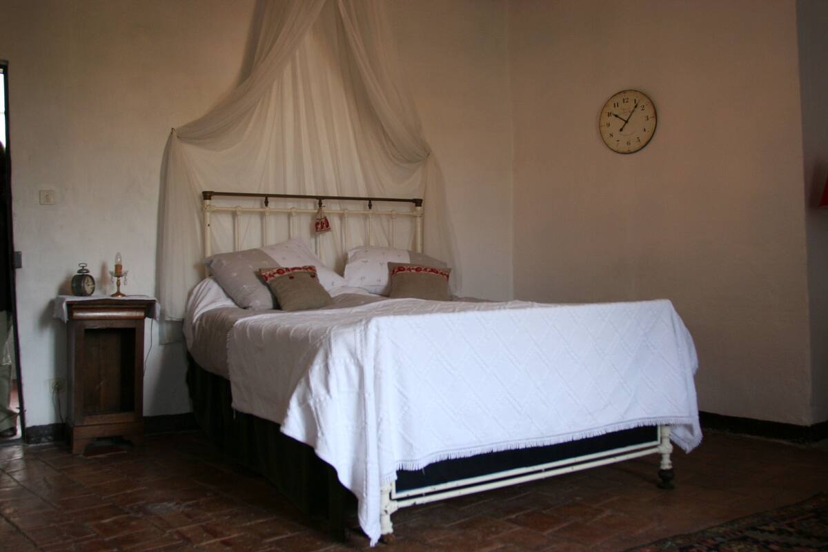 A bedroom with a four-poster bed, a bedside table with a lamp, and a wall clock.