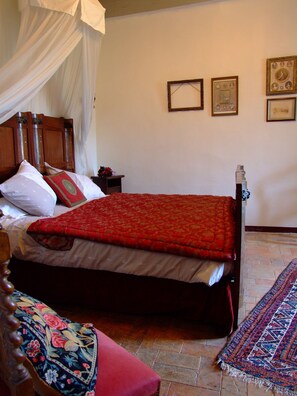 A bedroom with a wooden bed, a red bedspread, a canopy, a nightstand with a lamp, and a patterned rug. - Château de Rosans (Rosans)