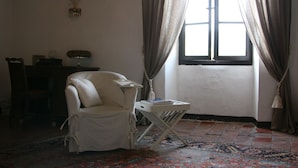 A room with a white armchair, a small table with a lamp, a window with curtains, and a patterned rug. - Château de Rosans (Rosans)