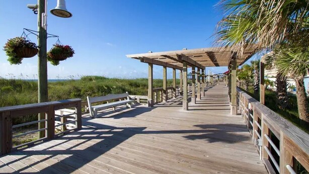 A wooden boardwalk with benches and hanging flower baskets, leading to a covered area with a view of the beach. - Ocean View Condo,  Steps to Beach & Boardwalk, 3BR/2BA! ★★★★★   (Carolina Beach)