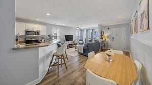 A modern kitchen with white cabinets, a microwave, and a wooden countertop. There is a dining area with a wooden table and white chairs. - Ocean View Condo,  Steps to Beach & Boardwalk, 3BR/2BA! ★★★★★   (Carolina Beach)