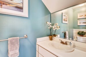 A bathroom with a blue wall, a white sink, a towel rack, and a mirror reflecting a plant and a picture frame. - Shared Pool + Gallery Art | Walk to Duval + Southernmost | Duval Sea Turtle (Key West)