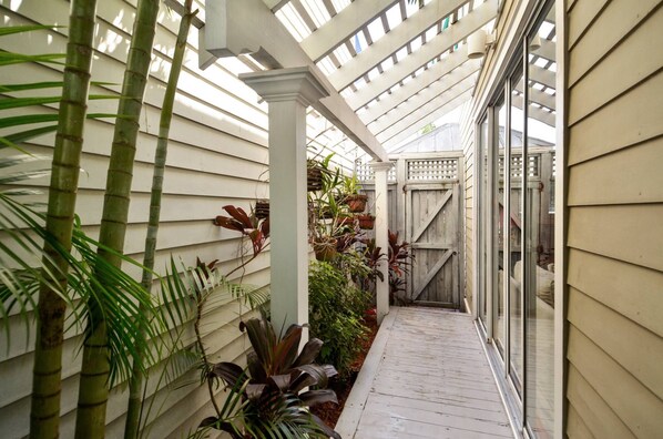 A covered walkway with bamboo plants, a wooden fence, and a glass door leading to a room.