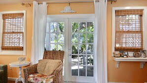 A room with a wicker chair, a small round table, and a view through a glass door to an outdoor area with plants. - Tranquility by AvantStay | Close to Duval St w/ BBQ & Shared Pool (Key West)