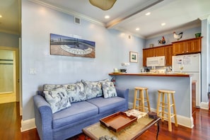 Sala de estar com sofá azul, mesa de centro de madeira e área de cozinha com armários de madeira e micro-ondas.