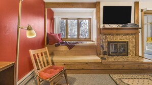 A cozy living room with a wooden bench, a red wall, a fireplace, and a large window. - RMR: Juniper 112 Condo in The Aspens/Wilson,WY (Wilson)