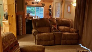 A cozy living room with a brown leather sofa, a patterned armchair, and a small table with a decorative item. A wooden door and window are visible in the background.