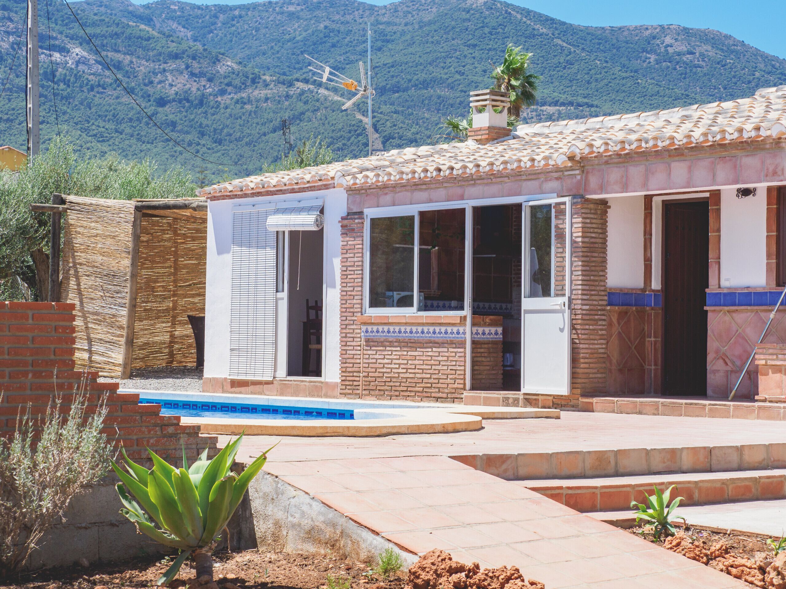 Un petit bâtiment avec une piscine, entouré d’un jardin planté de végétaux, et une montagne en arrière-plan.