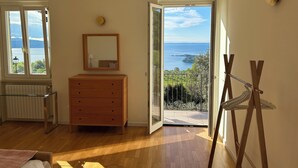 A bedroom with a wooden dresser, a mirror, and a balcony with a view of the sea.