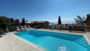 A rectangular swimming pool with a tiled deck, surrounded by lounge chairs and umbrellas, with a clear view of the sea and mountains in the background.