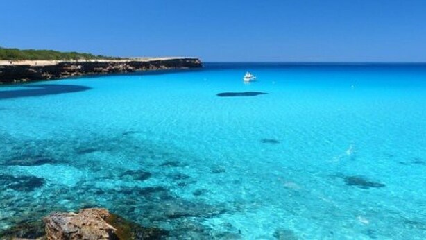 Un paysage côtier avec une eau turquoise et limpide, une côte rocheuse et un bateau au loin.