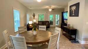 A dining area with a round wooden table, white chairs, and a vase with flowers. There is a ceiling fan, a television, and a framed picture on the wall. - Pet-Friendly Lakefront Retreat • Hot Tub, Sandy Shore & Fenced Yard (Westminster)