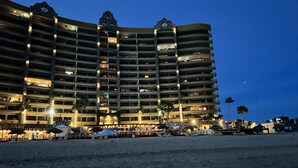 Un hotel a più piani con balconi, una spiaggia in primo piano e palme.