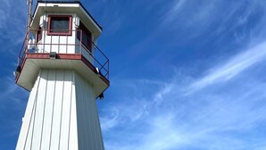 Ein weißer Leuchtturm mit einem roten Balkon, einer Holzterrasse und freiem Blick auf das Wasser.