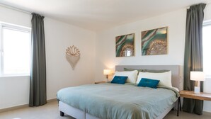A modern bedroom with a large bed, two wall-mounted artworks, and a bedside table with a lamp. - Fantastic ocean view apartment on Blue Bay (Sint Michiel)