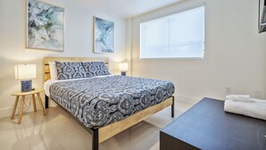 Une chambre moderne avec un grand lit, deux lampes de chevet, une table de nuit en bois, une grande fenêtre et deux tableaux abstraits accrochés au mur.