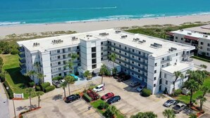 Un hotel de varios pisos con piscina en la azotea, situado cerca de una playa con vista al océano y gente disfrutando.