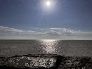 A clear view of the ocean with the sun reflecting off the water, a rocky shoreline in the foreground, and a partly cloudy sky. - Sea La Vie 4 min to Seawall or Cruise Port (Galveston)