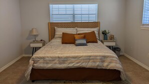 A bedroom with a large bed, a nightstand with a lamp, a small table with a plant, and a window with blinds.