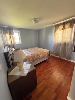 A bedroom with a bed, a bedside table with a lamp, a window with curtains, and a wooden floor. - The Paradise @ Lyncott, A famiily friendly place to stay! (Lansing)