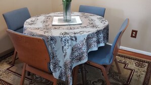 A round dining table with a patterned tablecloth, surrounded by four chairs with blue upholstery. A vase with white flowers is placed on the table.