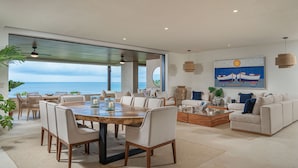 Un vaste espace de vie comprenant une grande table à manger, des sièges confortables, un canapé et une table basse. Une grande fenêtre offre une vue sur l’océan.