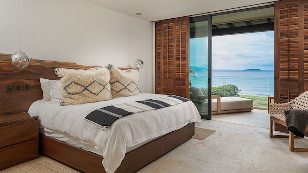 Une chambre avec un grand lit, une tête de lit en bois et une vue sur l’océan par une porte-fenêtre coulissante.