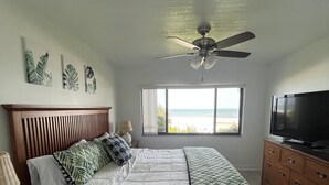 Een slaapkamer met een bed, een plafondventilator, een televisie en een raam met uitzicht op de oceaan.