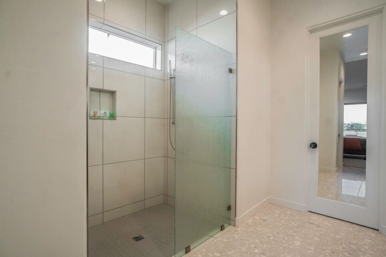 Une salle de bain moderne avec une douche à parois de verre, une petite étagère contenant des articles de toilette et une porte donnant sur une autre pièce.