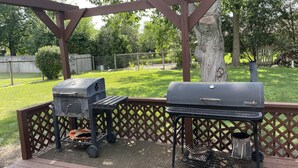 Ett takförsett utomhusområde med två grillar, ett picknickbord och ett trädäck.