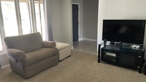 Ett vardagsrum med en beige soffa, en platt-TV placerad på ett glasbord och ett golv täckt av matta.