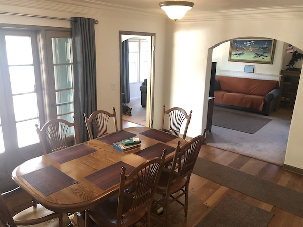 Uma sala de jantar com mesa e cadeiras de madeira, um cão ao fundo, e uma sala de estar com sofá e um quadro na parede.