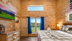 Un dormitorio de cabaña de madera con una cama, un aparador, una ventana con cortinas y un cuadro en la pared.